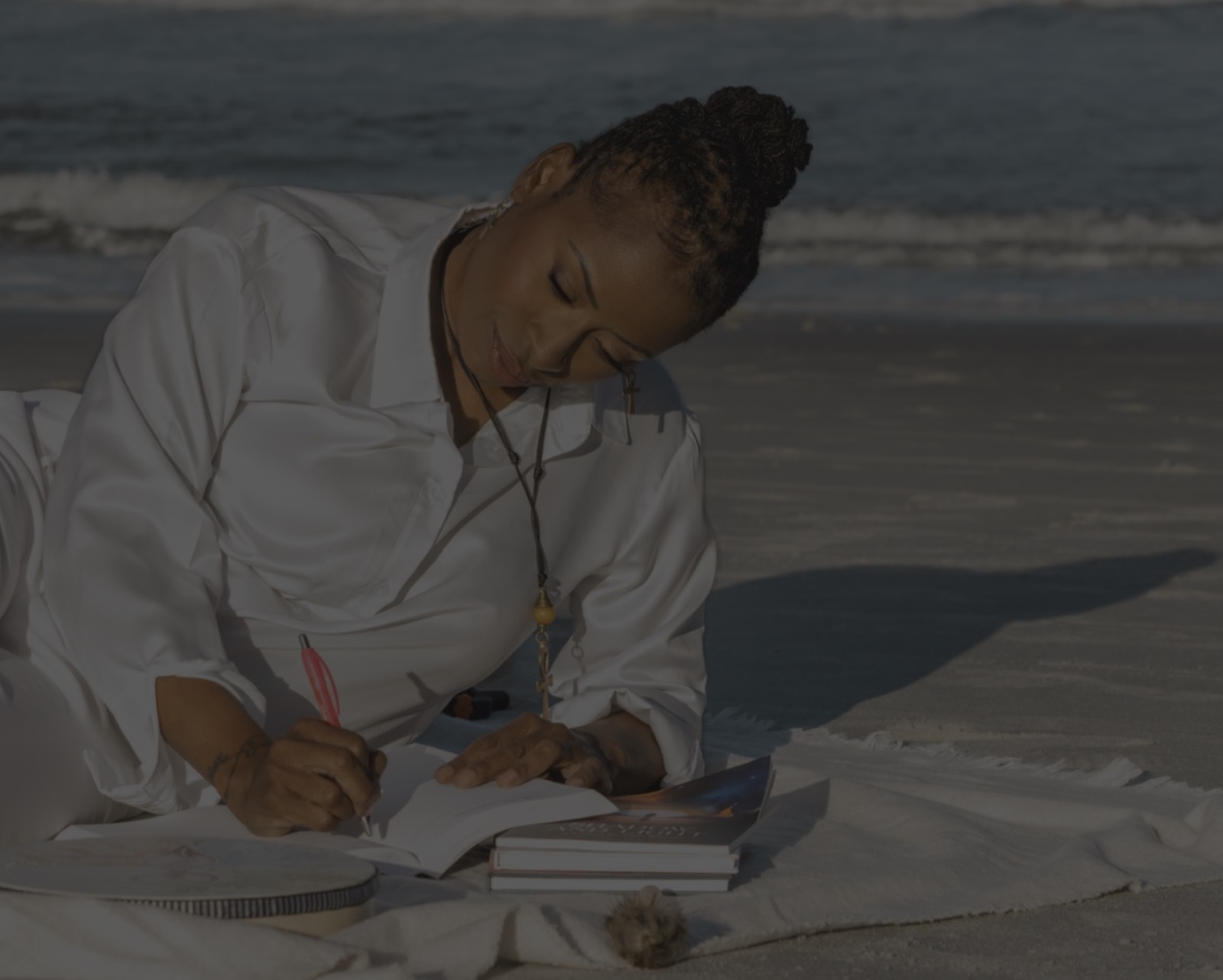 Latasha Smith journaling in the sand on the beach at sunset, with a stack of books beside her.